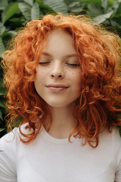 Portrait of a red head curly girl with heart shaped fake freckles staying in front of the leaves smiling with closed eyes