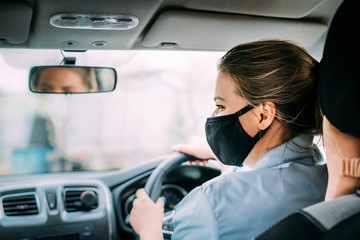 A young woman  taxi driver in a black medical mask with blond hair in a jacket rides a left-hand...