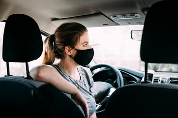 A young woman in a black medical mask with blond hair in a gray T-shirt is sitting in a left-hand drive car, put her hand on the seat. Portrait photo. Quarantine concept. Virus. Pandemic. Coronavirus