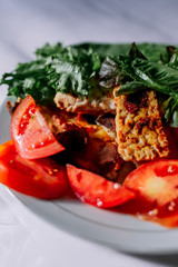 Traditional indonesian culinary food. Fried tempe in soy sauce, on red and black pepper. Greens and vegetables on a white plate. Beautiful sunlight is falling from the window.