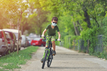 Happy kid having fun near home with a bicycle on beautiful spring day wearing protection mask for coronavirus Covid-19 pandemic virus