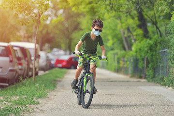 Happy kid having fun near home with a bicycle on beautiful spring day wearing protection mask for coronavirus Covid-19 pandemic virus