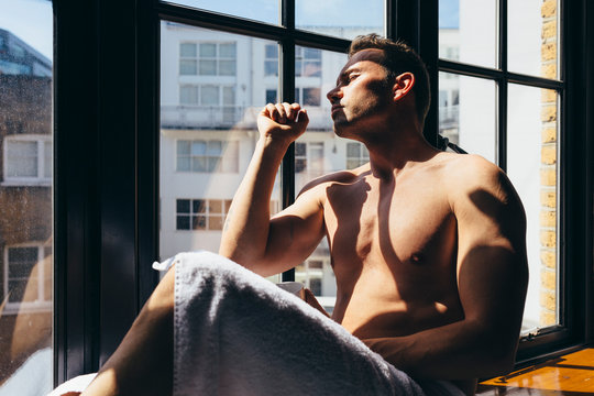 Sexy adult man enjoying the sun rays sitting besides the window