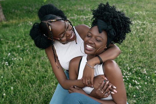 Two african girls in jeans and white tops are hugging on the grass in the park