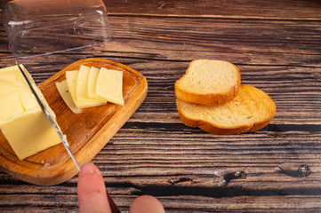Someone cuts some butter with a knife from a piece of butter in a wooden butter dish, slices of cheese and fresh wheat toast on a wooden background. Close up.