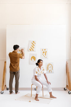 young man hanging paintings on wall while girl lounges in artist studio