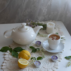 porcelain white teapot and white cup with tea on a wooden white table. candles, lemon and a bouquet of flowers