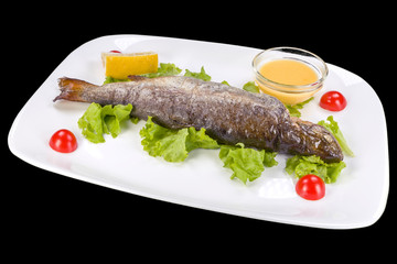 Tasty trout on lettuce served with sauce and lemon on a plate, isolated on black background