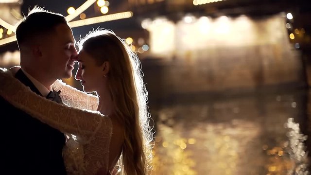 Portrait of young attractive couple in love, on night city background, lanterns, bridge and river, bokeh and blurred background. Bride and groom silhouette, slow motion, evening. backlight. Wedding