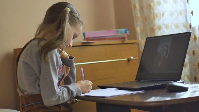 Little blond girl with two ponytails studies with her teacher that shows flashcards using video chat and write down something into exercise book at home because of the self-isolation due to Covid-19