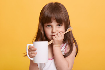 Closeup portrait of cute sad female child wearing t shirt, doing morning dental procedures, looks upset, having toothache, has problems with milk tooth, posing isolated over yellow studio background.