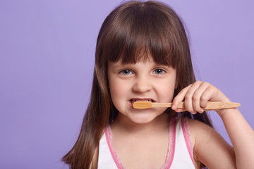 Close up portrait of caring sweet kid looking directly at camera, brushing teeth, keep clean, having straight hair, wearing shirt, posing isolated over lilac background in studio. Hygiene concept.