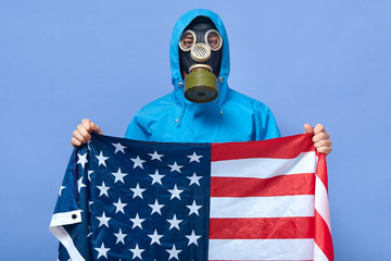Portrait of serious liquidating agent holding usa flag in hands, wearing gas mask and protection...