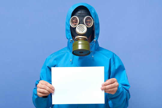 Indoor picture of man in protection uniform with gas mask on face holding blank sheet of paper, standing isolated over blue background in studio, being in danger. Copyspace for advertisement.