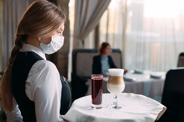 A female Waiter of European appearance in a medical mask serves Latte coffee.