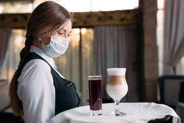 A female Waiter of European appearance in a medical mask serves Latte coffee.