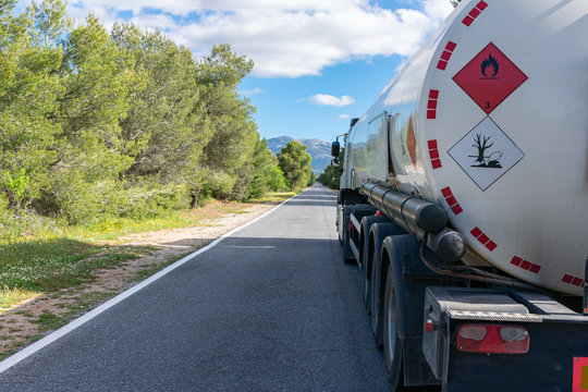 Hazard Labels For Flammable And Polluting Liquid On A Fuel Tank Truck Driving On A Narrow, Straight Road Lined With Trees.