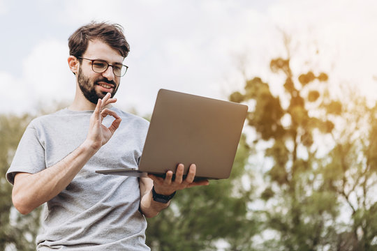 Stylish young man in glasses with laptop in hands in park on sky background working remotely connecting to wi fi
