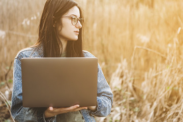 Happy hipster young woman working on laptop in field. Outdoors nature journey and relaxation. Freelance work concept.