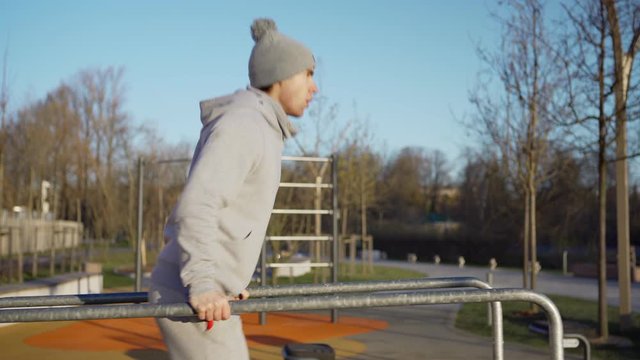 Tracking medium side view shot of athletic young man doing traveling dips exercise on parallel bars at street workout spot and recovering after