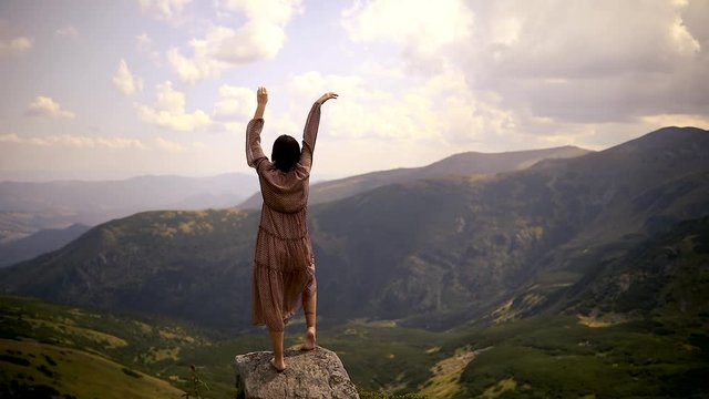 Camera moves around beautiful woman in light long dress lifting her hands up against sunset on mountain top. Girl in a long dress that flutters in the wind