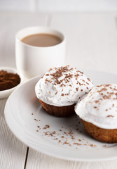 Cupcakes with whipped cream and chocolate chips on a white plate and a Cup of coffee in the background, on a white wooden table. Image for the menu or catalog of confectionery products.