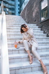 The beautiful caucasian woman holds the juice in her hand and sits on the staircase in front of the building