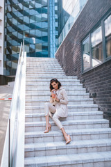 The beautiful caucasian woman holds the juice in her hand and sits on the staircase in front of the building