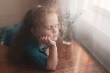 Sad little girl sadly looks out the window. Shooting through the glass. Selective focus