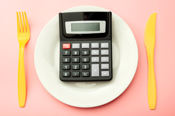 Calculate expensive food spending costs, counting calories and weight loss program concept with calculator onn empty plate, yellow fork and knife isolated on pink background