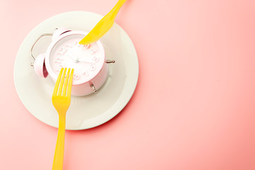 Weight loss strategy metaphor and intermittent fasting plan concept with minimal photography of clock on empty plate and vibrant yellow fork and knife isolated on vivid pink background with copy space