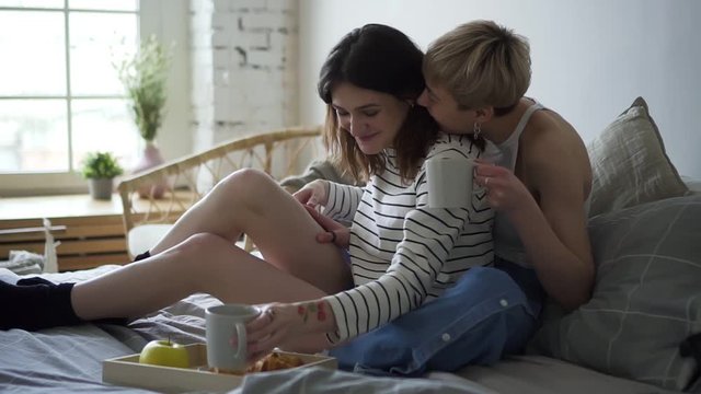 lesbian couple drinking coffee while sitting on bed. they talk, smaile and laugh, Spbd concept tolerance, same sex, love. pretty women communicate and relax at home.