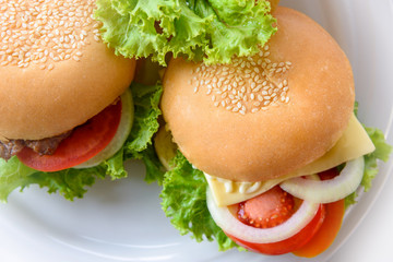 Closeup top view two cheeseburger made from pork or beef, green lettuce bread onion white sesame and tomato on white plate at table background made at home. Hamburger is delicious American fast food