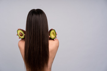 Brunette with long hair stands with his back to the camera, she's naked shoulders, and in the hands of the avocado halves, concept health and beauty