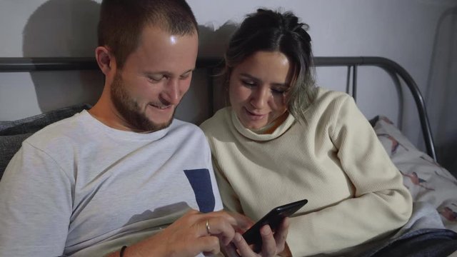 Happy Young Couple Use Smartphone in Bed at Night, They Share Screen, Browsing Through Social Networks, Sharing Pictures, Using Internet and Having Fun. Top View Camera Shot with Blue Colors