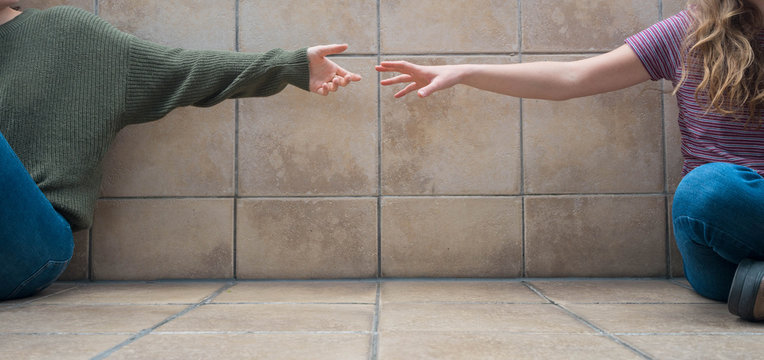 Social Distance. Front View Of Two Girls Sitting On The Floor. They're Spaced Out And Trying To Put Their Hands Together