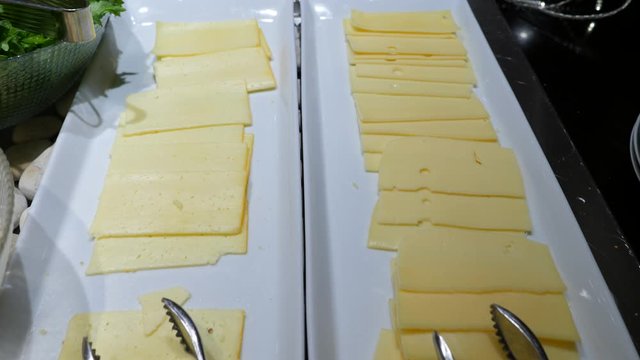 Two long plates with sliced cheese at buffet breakfast, cold starters section, stainless steel tongs lie for self service.