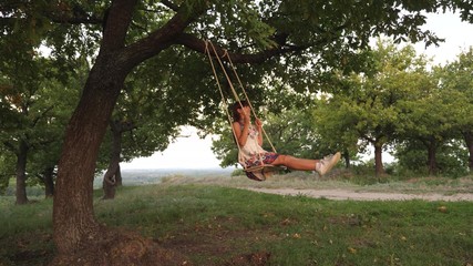free young girl swinging on rope swinging on an oak branch. healthy child swinging on swing in park in sun. Girl loves to fly on a swing in the forest. concept of happy family and childhood.