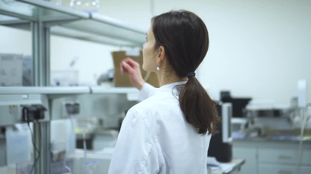 Young woman lab assistant is walking in interior of pharmaceutical laboratory spbd. American female professional is taking glass flask from shelf during working day in modern company. Employee wearing