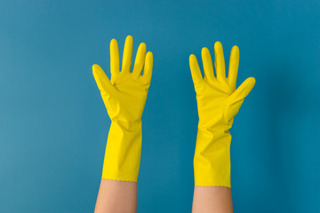 Close up on hand of unknown caucasian woman female girl holding up hands with protective yellow rubber gloves used to to clean or disinfection against blue wall background in day