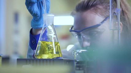 female chemist pour liquid in flask with pipette. Spbd Yellow liquid is mixed in a glass beaker in a laboratory. color change in laboratory. concept research, analysis, experiment. reaction on stirrer - Powered by Adobe