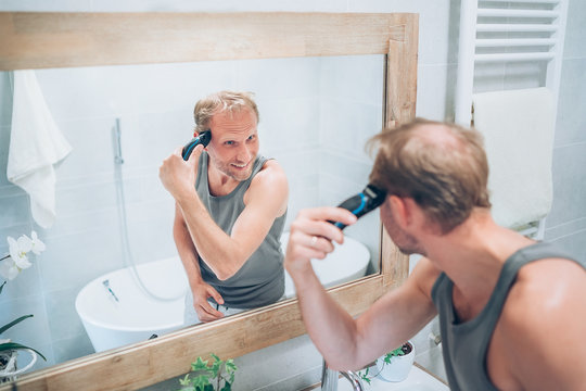 Smiling Man making new style haircut trimming a hairs  using an electric rechargeable Trimmer looking in bathroom mirror. Hairstyle, Body and skincare treatment concept.