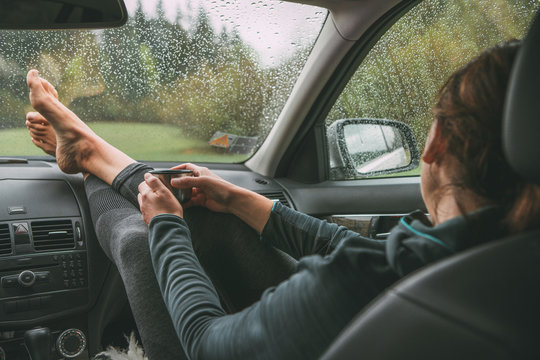 Female holding the hot tea tourist thermos mug. She sitting on co-driver seat inside modern car, putting her legs on car dashboard, enjoying a moody rainy day weather looking through windscreen drops