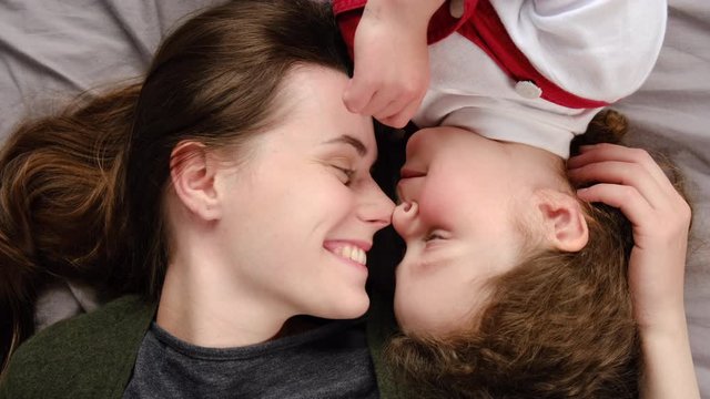 Top view of happy young mother and little child daughter playing bonding touching noses laughing together lying in bed at home, cheerful affectionate mom having fun with cute small kid enjoy cuddling 