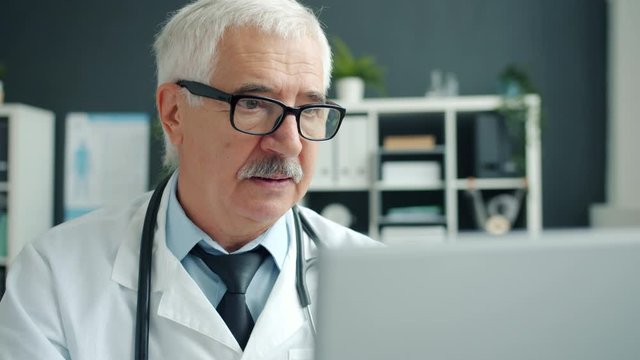 Serious doctor in white gown is using computer then touching glasses working alone in hospital. Online consultation, people and profession concept.