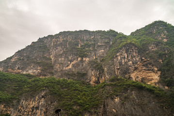 Fototapeta na wymiar Wuchan, China - May 7, 2010: Dawu or Misty Gorge on Daning River. Tall huge mountains with flanks like cliffs and some green foliage on top towering over canyon under gray cloudscape.
