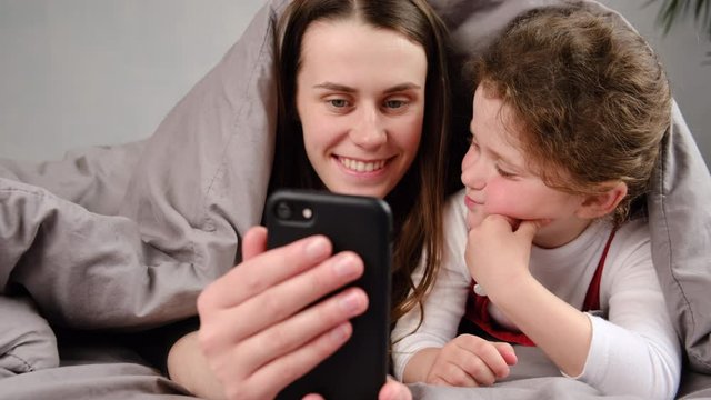 Happy young woman lying on bed covered with blanket with cute smiling little kid daughter, making selfie photo on smartphone. Joyful family of two using mobile applications together in bedroom