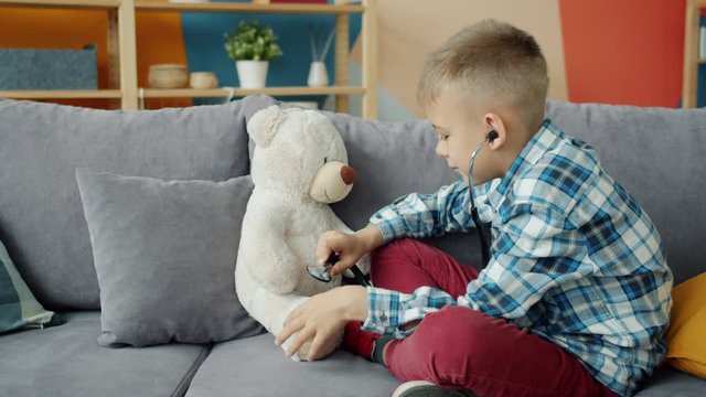 Cute kid is playing doctor with teddy bear using stethoscope smiling having fun sitting on sofa indoors at home. Childhood, creative games and people concept.