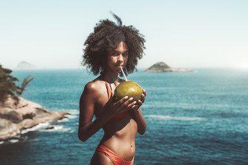 Young foxy Brazilian female¬†with curly afro hair and in swimsuit is standing on an island beach with the ocean in the background, holding coconut in her hands and drinking coco water using a straw - Powered by Adobe