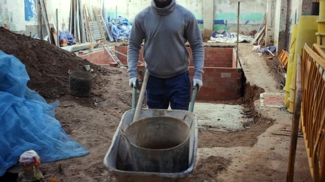 Young bearded man working at building site, carrying navvy wheelbarrow with bucket of construction mortar